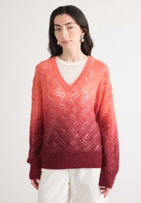 Pull en maille à col en V avec un dégradé de corail à bourgogne. Présente un motif texturé et des manches longues, superposé sur un haut blanc.