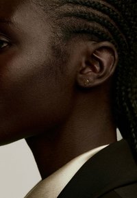 Pendiente en forma de estrella dorada sobre piel oscura, con una vista cercana de la piel texturizada y el cabello trenzado. Tela negra y crema visible en el fondo.