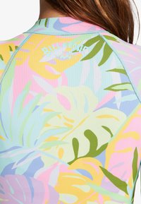 Camisa de protección multicolor con un patrón floral en tonos suaves de rosa, azul, amarillo y verde, con un detalle de costura azul y logo.