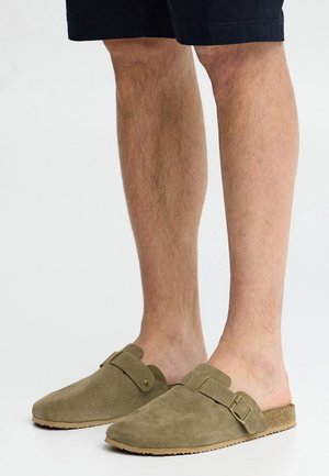 Beine in schwarzen Shorts und olivgrünen Wildleder-Slippern mit seitlicher Schnalle, stehend vor einem schlichten weißen Hintergrund.