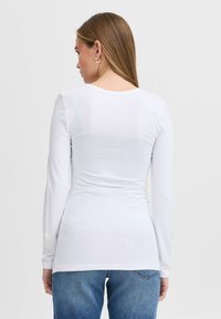 Long-sleeve white top with a fitted design and round neckline, made of smooth fabric. Back features subtle ruching and is paired with blue jeans.