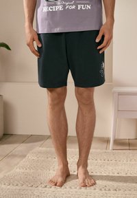 Mörkgröna shorts i bomull med en broderad tecknad design. Tyget har en mjuk yta och passformen verkar avslappnad.