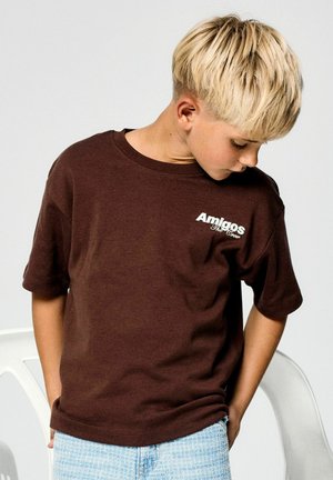 Blond jongetje draagt een bruin "Amigos The Crew" t-shirt en lichtblauwe short, kijkt naar beneden met handen in de zakken tegen een effen achtergrond.
