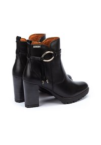Botas negras de cuero hasta el tobillo con punta redonda, un tacón grueso y texturizado, y un acento metálico circular cerca del tobillo. Interior forrado en naranja.