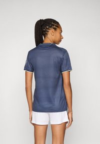 Marinblå atletisk tröja med korta ärmar, texturerad tyg, och rundad halsringning, kombinerad med vita shorts. Bakifrån syns flätat hår.