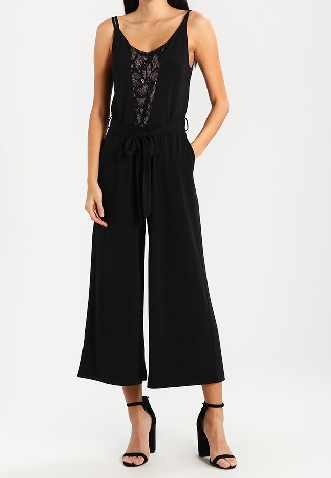 Femme portant une combinaison noire sans manches avec un panneau avant en dentelle et un pantalon jambes larges, associée à des sandales noires à talons hauts.