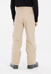 Pantaloni impermeabili beige con una texture liscia, gamba ampia e vita regolabile, abbinati a robusti stivali neri su uno sfondo bianco pulito.