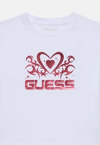 Weißes Baumwoll-T-Shirt mit einem roten Folienherz-Graphic und dem "GUESS"-Logo darunter, verziert mit geschwungenen Mustern und diamantähnlichen Akzenten.