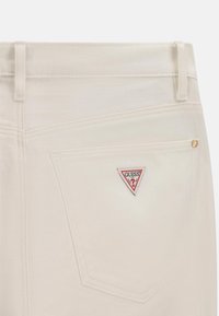 Poche arrière d'un jean en denim blanc présentant un patch logo Guess triangulaire rouge et blanc et un rivet doré dans le coin supérieur droit.