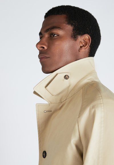 Manteau beige avec un col haut, présentant un détail de bouton et une texture lisse. Le design comprend des accents de couture subtils.