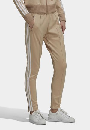 Persoon die beige trainingsbroek met witte strepen en ritszakken draagt, gecombineerd met witte sneakers, staand met handen in de zakken.
