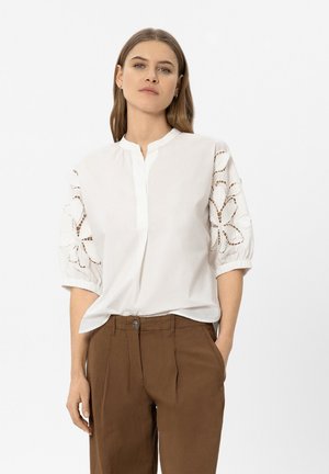 Woman wearing white blouse with cut-out floral sleeves and brown high-waist pants, standing against plain light background.