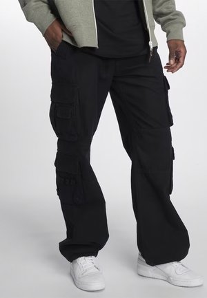 Personne portant un pantalon cargo noir à multiples poches, des baskets blanches, une veste grise à fermeture éclair, et un t-shirt noir, debout avec une main dans la poche.