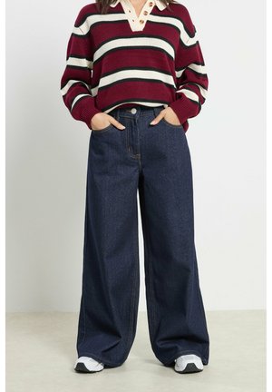 Persoon die donkerblauwe jeans met wijde pijpen, witte sneakers en een rood-wit-zwart gestreepte trui met knoopkraag draagt, handen in de zakken.