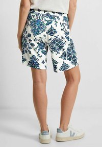 Pantalones cortos blancos con un patrón de flores azules, cinturilla elástica y bolsillos laterales. El modelo lleva zapatillas beige con detalles en azul claro.