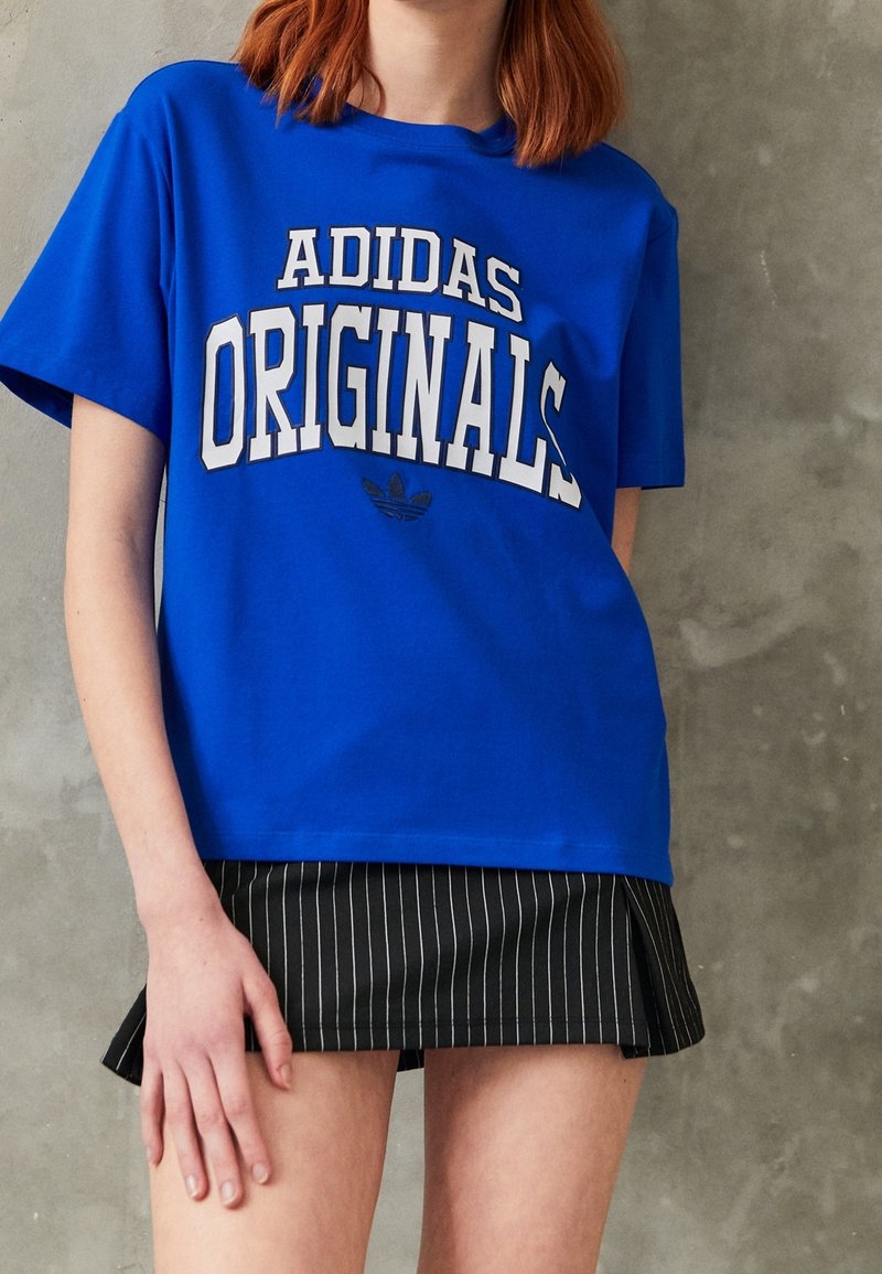 Osoba na sobě má modré tričko Adidas Originals a černou mini sukni s jemnými proužky, pozadí je obyčejná šedá zeď.