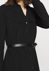 Robe noire boutonnée avec manches longues, deux poches poitrine et ceinture noire avec boucle circulaire dorée. Tissu lisse et coupe ajustée.