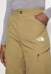 Homme portant un short beige de North Face avec une poche latérale zippée, la main dans la poche, et un t-shirt à manches longues gris.