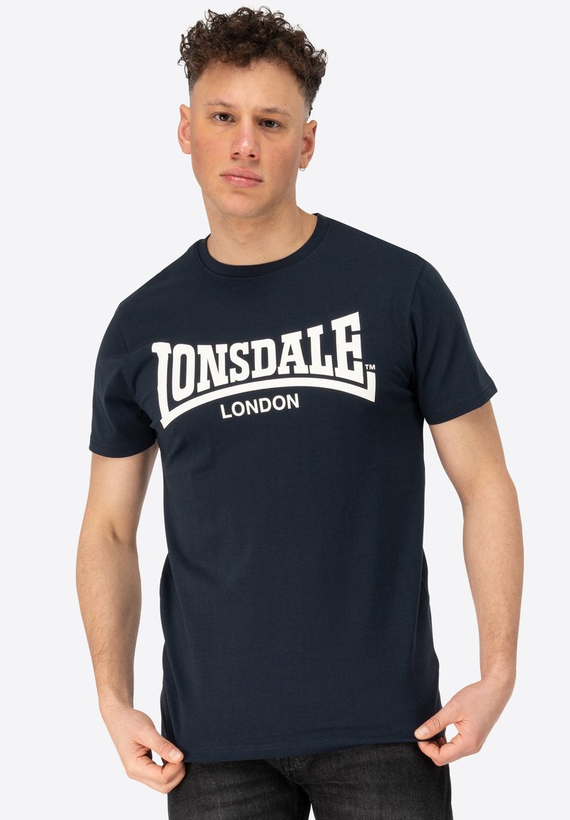 Tengerészkék pamut póló, a mellkasán nagy, fehér nyomott "LONSDALE LONDON" logóval. Rövid ujjú és kerek nyakú kialakítás.