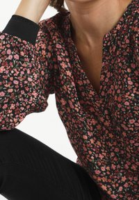 Zwarte blouse met bloemenpatroon, versierd met roze en groene bloemen. De blouse is gemaakt van een zachte stof en heeft een knoopsluiting aan de voorkant, evenals gathered mouwen met geribbelde manchetten.