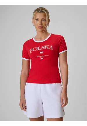 Kobieta ubrana w czerwoną koszulkę z napisem "POLSKA" oraz białe szorty, stojąca na jednolitym szarym tle, zwrócona przodem.