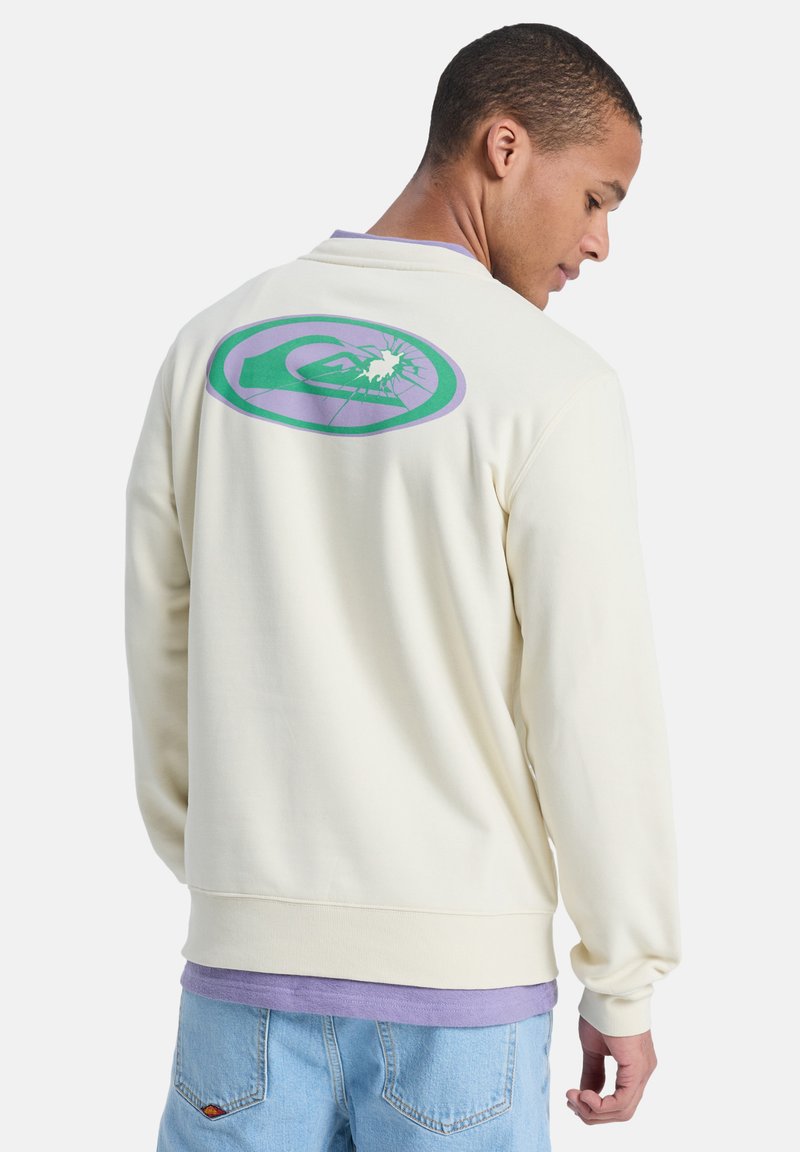 Sweatshirt de couleur crème avec un motif rond en violet et vert représentant une montagne au dos. Texture douce et coupe décontractée.