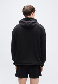 Μαύρο φούτερ με κουκούλα και λεία υφή, με άνετη γραμμή και μακριά μανίκια, σε προβολή από πίσω. Φορεμένο με μαύρο σορτς.