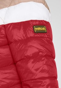 Úsek rukávu červené, bílé a béžové podšívkové bundy se žlutou a černou nášivkou loga Barbour International.