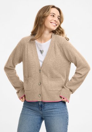 Frau lächelt, trägt eine beige Strickjacke mit Knöpfen und Taschen über einem weißen Hemd und blauen Jeans, steht mit Händen in den Hüften.