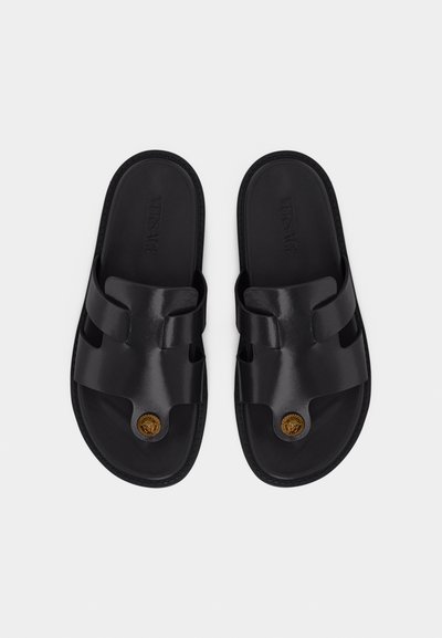 Μαύρα δερμάτινα σανδάλια slide με φαρδιές λουριά, εσωτερικά πατάκια με ανάγλυφο το λογότυπο "Versace" και χρυσό έμβλημα Medusa στην περιοχή των δαχτύλων, εμφανιζόμενα από πάνω.