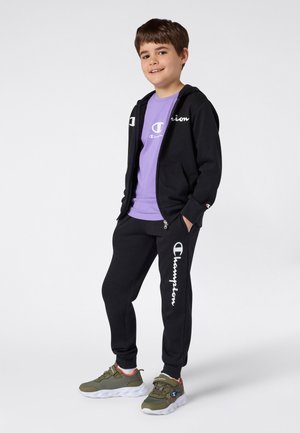 Felpa nera con zip e pantaloni, t-shirt viola sotto, con dettagli del logo bianchi; scarpe sportive verdi e grigie con accenti arancioni.