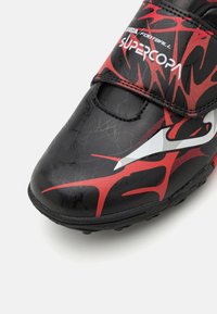 Botín de fútbol negro con acentos en rojo y blanco, que presenta una textura agrietada y una correa de Velcro para ajustar el calce.