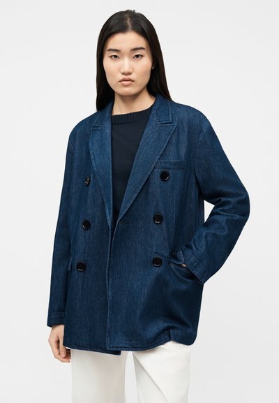 Γυναίκα που φοράει μπλε σκούρο διπλό μπλέιζερ από τζιν πάνω από μαύρο τοπ και λευκό παντελόνι, με το ένα χέρι στην τσέπη.