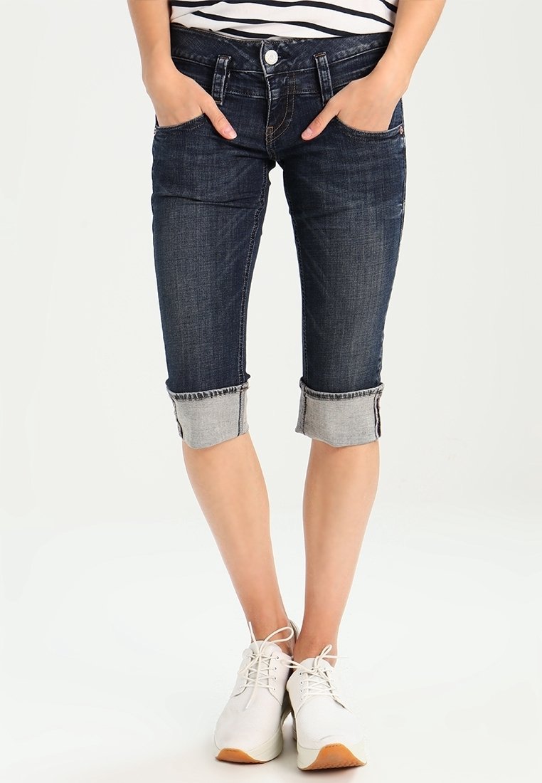 Tummat farkku capri-housut, joissa on käännettylahkeet, viisi taskua ja etunappi, yhdistettynä valkoisiin urheilukenkiin.