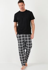 Camicia nera in cotone a maniche corte con tre bottoni, abbinata a pantaloni da pijama a scacchi bianchi e neri con vestibilità ampia e cintura elastica.