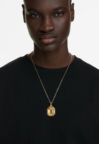 Guldtonet pendantsmykke med en ottekantet form, prydet med detaljerede facetter, hængende fra en delikat kæde, båret over en sort skjorte.