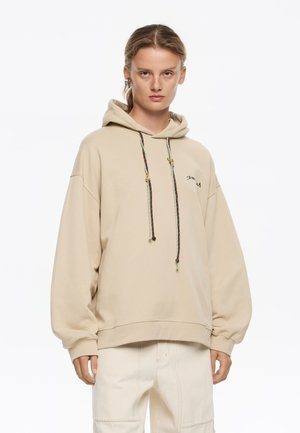 Sudadera beige de gran tamaño hecha de una tela suave con capucha ajustable, que presenta cordones decorativos multicolores y un pequeño logo bordado.