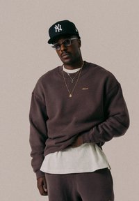 Brauner Sweatshirt mit Rundhalsausschnitt, versehen mit einem kleinen goldenen Logo. Getragen über einer weißen Rippenschicht; ergänzt durch eine schwarze Kappe und geschichtete goldene Halsketten.