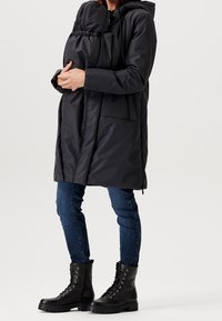 Abrigo de plumas negro con capucha, que cuenta con un bolsillo delantero y un panel elástico para llevar a un niño. Combinado con jeans oscuros y botas negras.