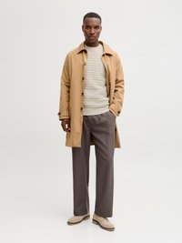 Uomo che indossa un trench beige, maglione grigio chiaro testurizzato, pantaloni grigio scuro e scarpe in suede beige, in piedi contro uno sfondo chiaro e semplice.