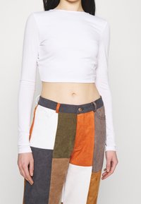 Weißes langärmeliges Crop-Top aus glattem Stoff, kombiniert mit gemusterten Cordhosen in mehreren Farben, die orange, grün und braun Töne zeigen.