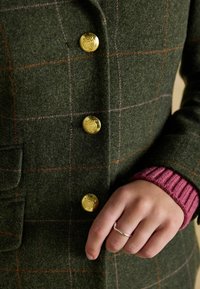 Vihreä villatakki, jossa on oranssi ja vaaleanpunainen ruutukuosi, kultanapit ja hopeasormusta pitävä käsi, kerroksittain vaaleanpunaisen ribbineuleen päällä.