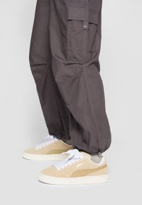 Pantalons cargo gris avec une coupe décontractée, dotés de poches latérales et de poignets élastiques, associés à des baskets en daim beige et crème avec des lacets blancs.