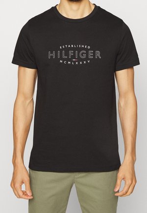 Mann trägt ein schwarzes kurzärmeliges T-Shirt mit dem Aufdruck "Established Hilfiger MCMLXXXV" und einem kleinen Design der rot-weiß-blauen Flagge auf der Vorderseite.