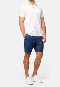 Chemise blanche en lin à manches courtes avec col, associée à un short bleu marine retroussé et des baskets blanches, mettant en avant un style estival décontracté.