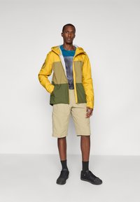 Farbblockierte Jacke mit gelben, grünen und grauen Abschnitten; verfügt über eine Kapuze und einen Reißverschluss. Getragen über einem blauen Shirt, kombiniert mit beigen Shorts.