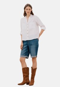 Camicia bianca in lino con bottoni e due tasche sul petto, abbinata a pantaloni shorts in denim blu. Stivali in suede marroni al ginocchio completano l'outfit.