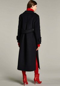 Lungo cappotto nero in lana con vita aderente, polsini abbottonati e fessure laterali, abbinato a un dolcevita rosso a coste e stivaletti rossi alla caviglia.