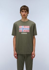 Camiseta de algodón verde oliva con un diseño abstracto impreso en rojo y azul que recuerda a la bandera noruega, y letras en negrita debajo.