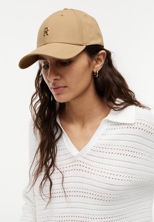 Frau mit langen braunen Haaren, die eine beige Kappe mit Initialen und ein weißes gestricktes Oberteil mit Kragen trägt, schaut nach unten vor einem schlichten Hintergrund.